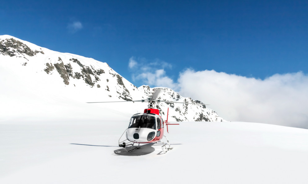 A helicopter rests on snow, with a large mountain range in the background